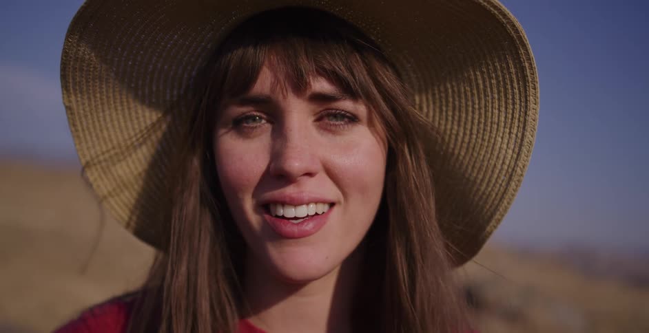 An expressive close-up of a young woman in a sun hat, her mood shifting from a bright smile to a look of worry in a sun-drenched landscape