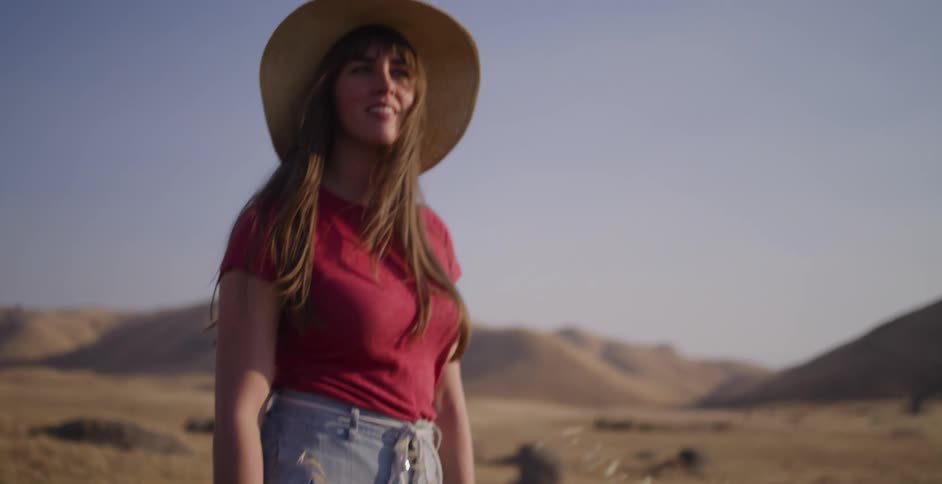 A woman in a straw hat stands in a sun-drenched, golden field with rolling hills in the background, enjoying the moment in slow motion