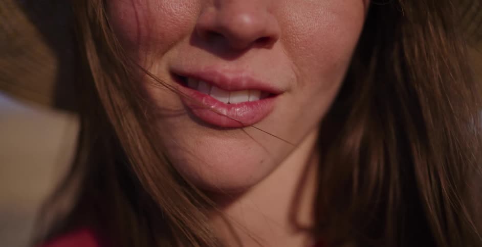An intimate slow-motion close-up of a young woman in a straw hat smiling softly as the wind blows through her hair