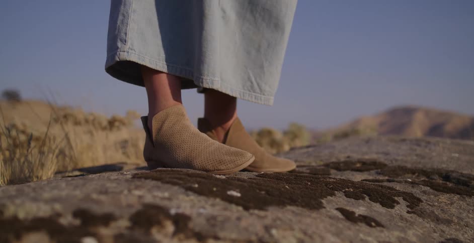 A woman wearing tan suede booties stands on a moss-covered rock in a sunlit, arid landscape