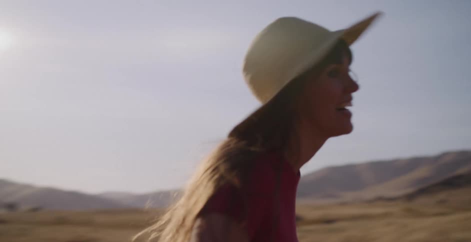 A joyful woman in a straw hat runs carefree through a sun-drenched, hilly landscape in slow motion