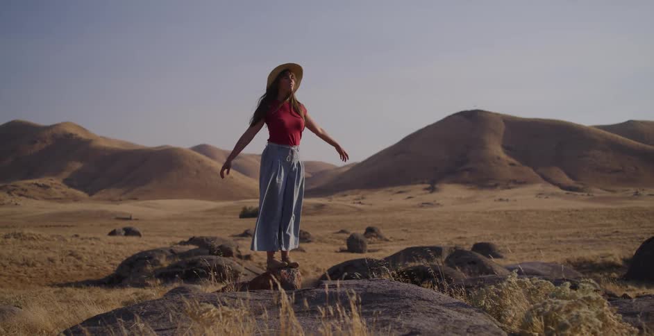 A young woman wearing a hat finds her balance on a large rock in a vast, hilly landscape, opening her arms to the sky in a moment of serenity