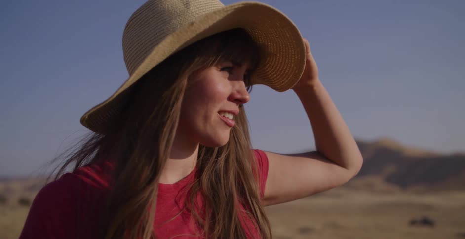A happy young woman in a wide-brimmed hat smiles as she looks out over a sunny desert landscape in slow motion