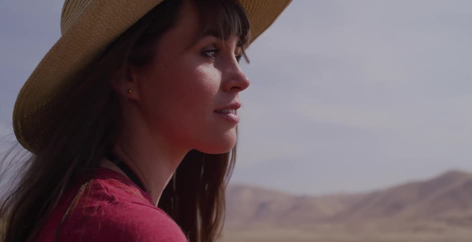A serene woman in a wide-brimmed hat closes her eyes to soak up the sun's warmth in a vast, golden, hilly landscape