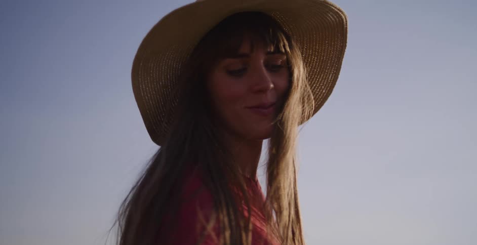 A woman in a sun hat gently touches dry grass while walking through a vast, arid field in slow motion