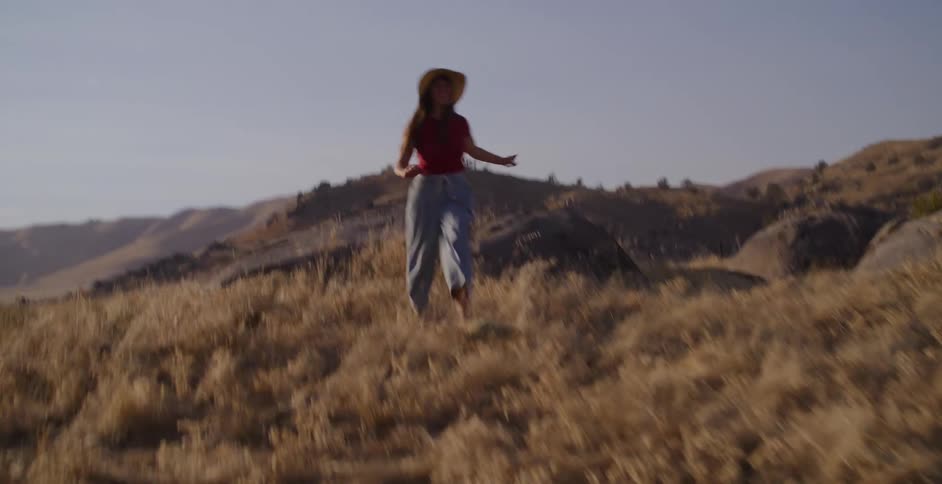 A carefree woman in a straw hat runs and dances joyfully through a golden grassy field in the hills on a sunny day