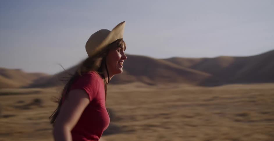 A joyful woman wearing a straw hat runs freely through a golden, sunlit field in slow motion