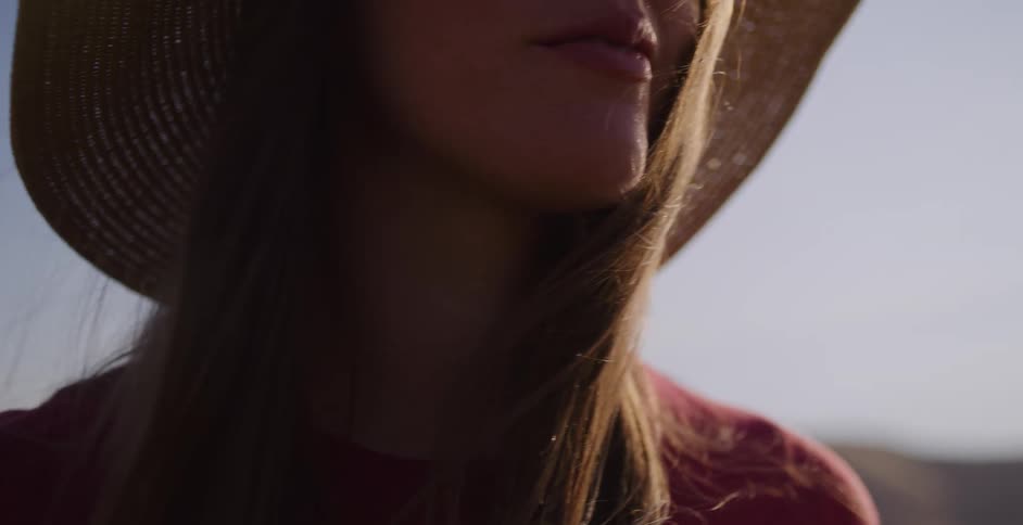 Close up of a thoughtful woman in a straw hat with her hair blowing in the wind during a serene golden hour