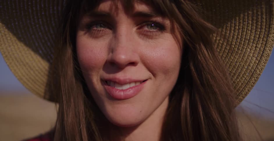 A slow-motion close-up of a beautiful young woman wearing a straw hat as she opens her eyes and smiles warmly at the camera