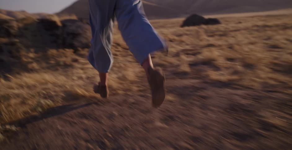 A woman wearing a hat and boots runs through a dry, sun-drenched landscape towards rolling hills in slow motion