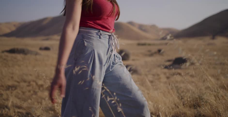 A woman wearing a straw hat walks through a golden field, gently touching the tall grass as the camera follows in slow motion