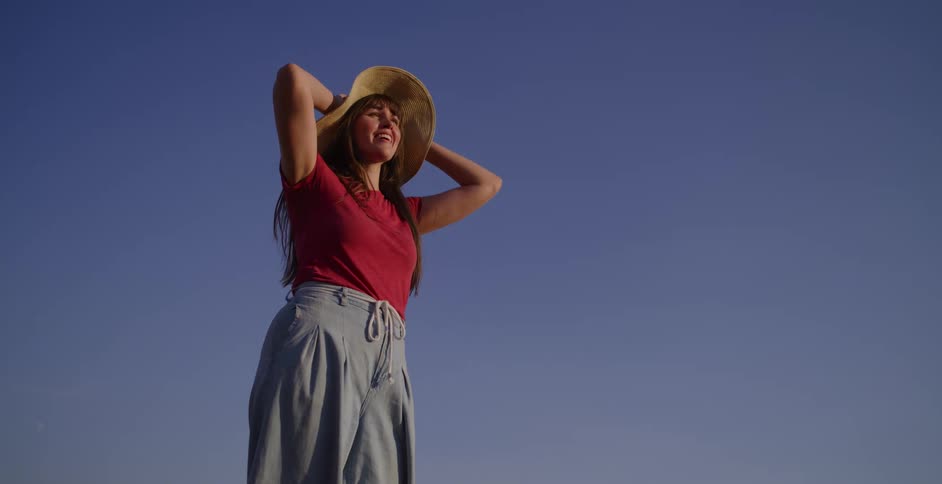 A low angle shot of a happy woman in a straw hat smiling and stretching against a clear blue sky