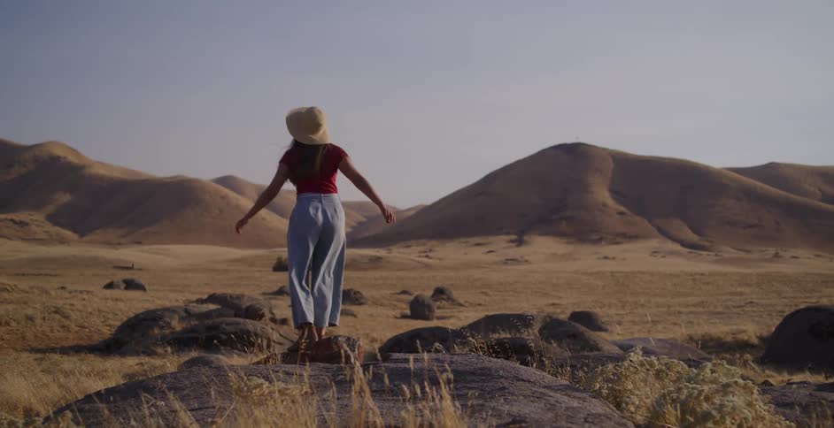 A woman wearing a straw hat stands atop a rock and stretches her arms out, taking in the vast, golden, hilly landscape at sunset