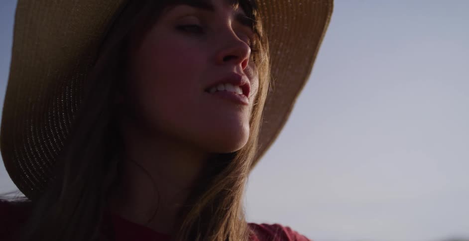 A serene woman in a straw hat closes her eyes and smiles, basking in the warm glow of the setting sun