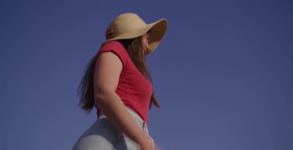 A slow tilt-up from boots on a mossy rock reveals a smiling woman in a straw hat standing in a sunny, arid landscape