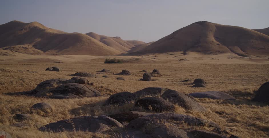 A vast, desolate landscape of rolling, golden hills and rocky outcrops under a hazy sky