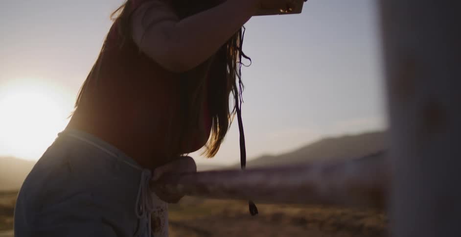 A woman in a straw hat leans against a rustic fence and takes a photo with an instant camera in slow motion at sunset