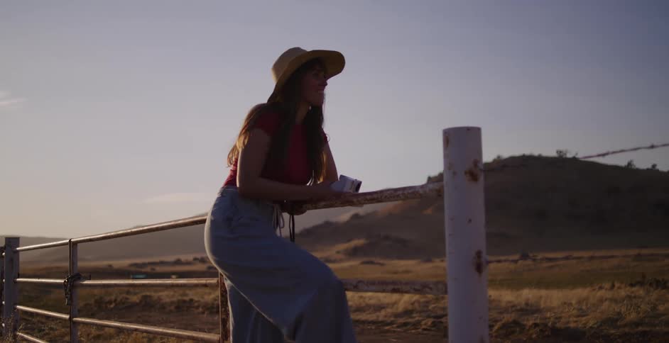 A young woman on a country road trip climbs a rustic fence at sunset to take a picture with her instant camera