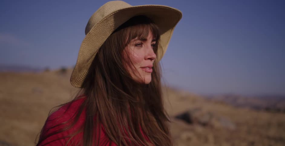 A beautiful woman in a straw hat smiles as she enjoys the sunshine and wind in a golden, hilly landscape