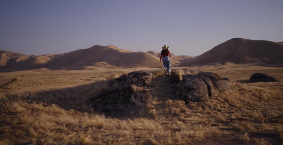 A free-spirited woman runs through a golden, hilly landscape, stopping on a rock and spreading her arms in a moment of joy and freedom
