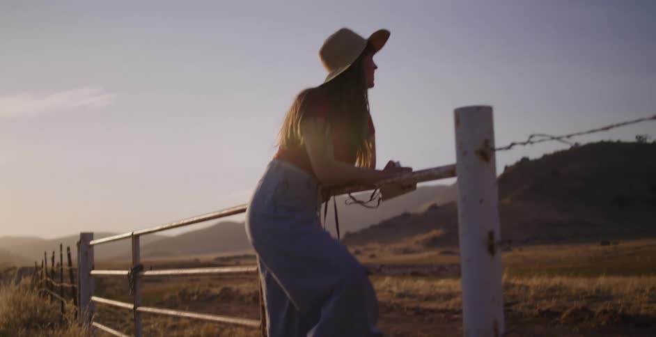 A free-spirited woman jumps out of her vintage car, hops a rustic fence in the country, and takes photos of the scenic landscape at golden hour