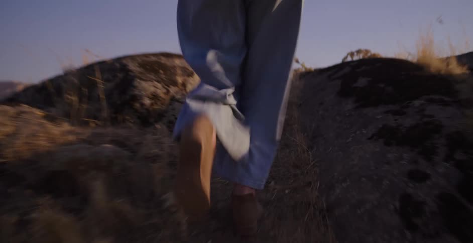 Following a woman's feet in boots as she runs up a rocky, grassy hill to overlook the landscape at golden hour