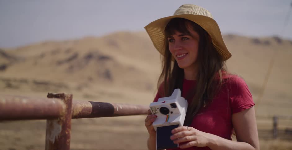 A smiling young woman in a sun hat captures a memory with her instant camera in a dry, rural landscape