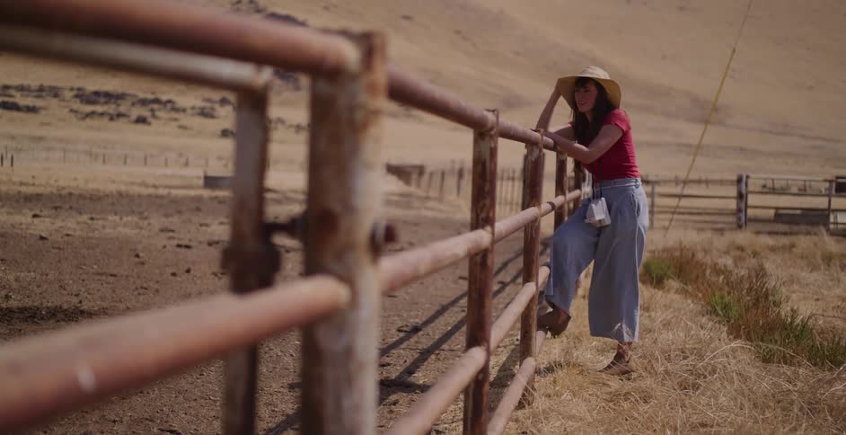 A thoughtful young woman in a sun hat leans on a rusty fence, looking over a dry, empty ranch corral on a sunny day