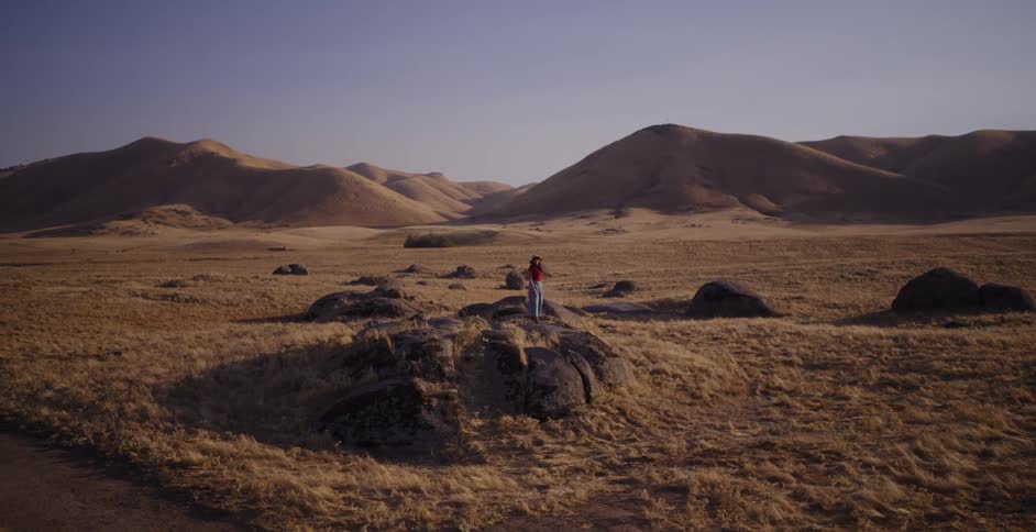 A carefree woman wearing a hat dances on a large rock in a vast, arid landscape with rolling golden hills in the background