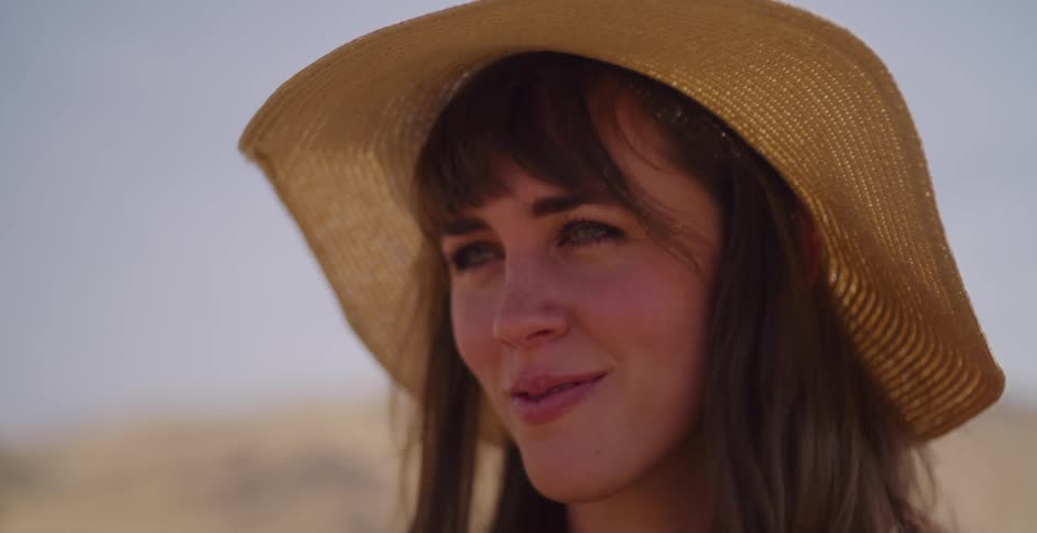 A slow-motion close-up reveals a serene woman in a straw hat looking thoughtfully into the distance in a sunny desert landscape