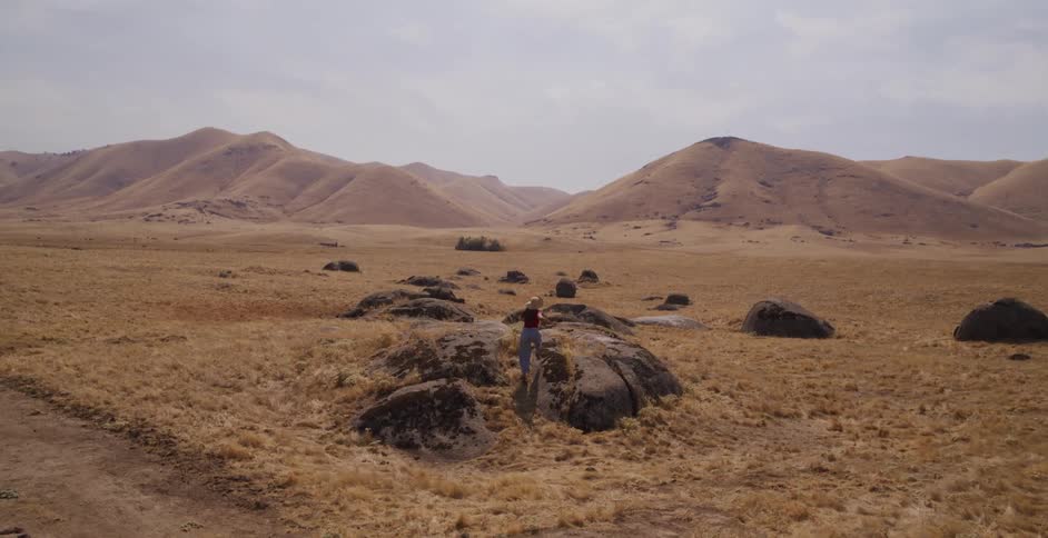A woman joyfully runs through a vast, arid landscape, climbs onto a large rock, and stretches her arms wide in a gesture of freedom