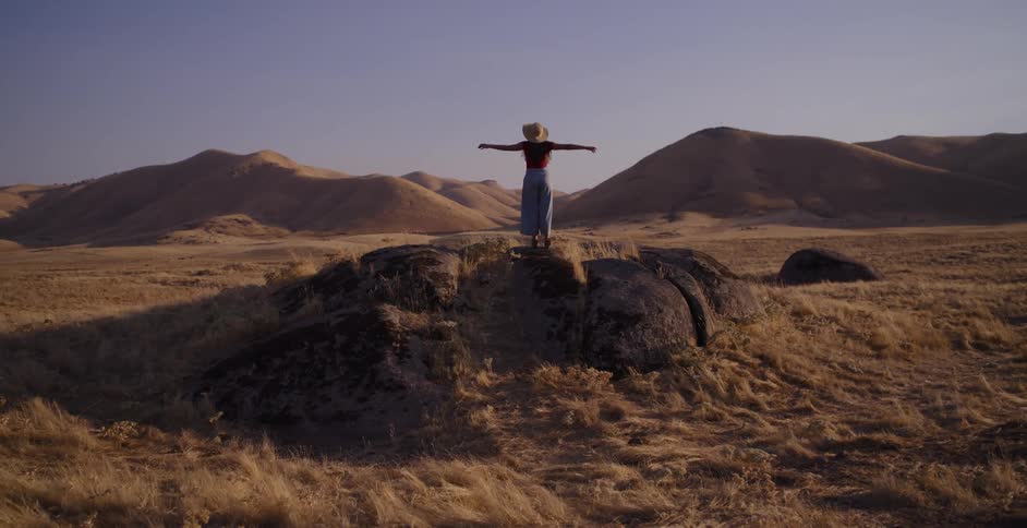 A free-spirited woman runs through a golden valley at sunset, climbing a rock and spreading her arms wide to embrace the scenery