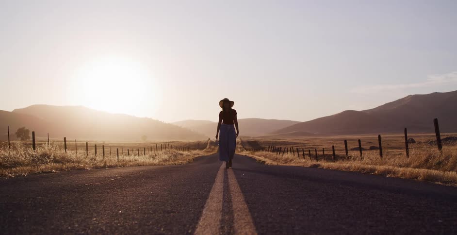 A stylish woman in a hat walks down the middle of a deserted country road towards the camera as the sun sets
