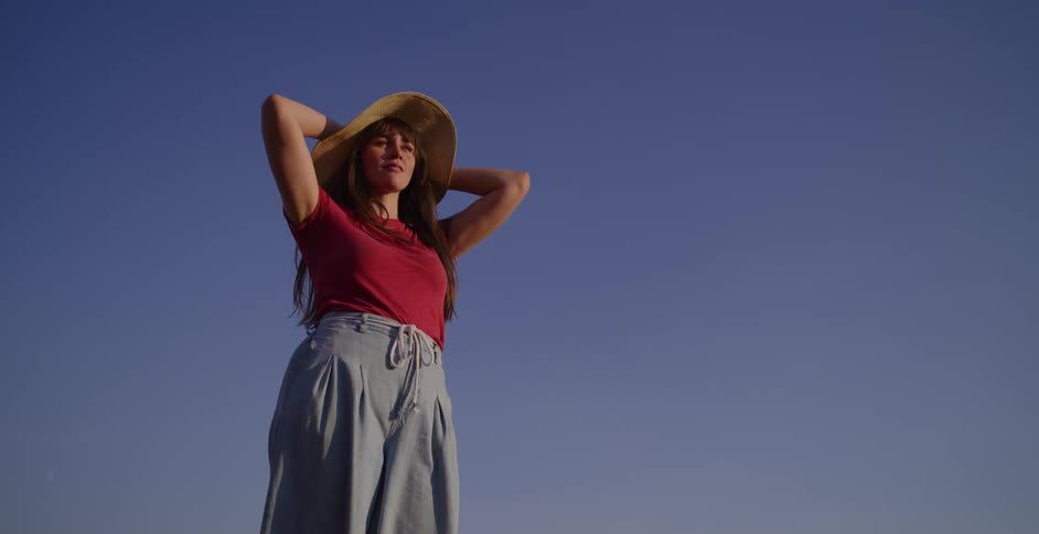 A happy woman in a straw hat stretches and smiles while enjoying the golden hour light against a clear blue sky