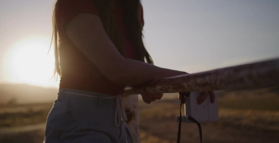 A young woman in a sun hat smiles while leaning on a rustic fence and raises her instant camera to capture the golden hour sunset
