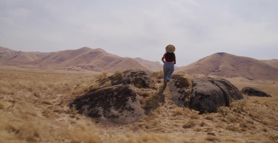 A free-spirited woman runs through a vast, dry landscape and stands triumphantly on a rock with her arms wide open