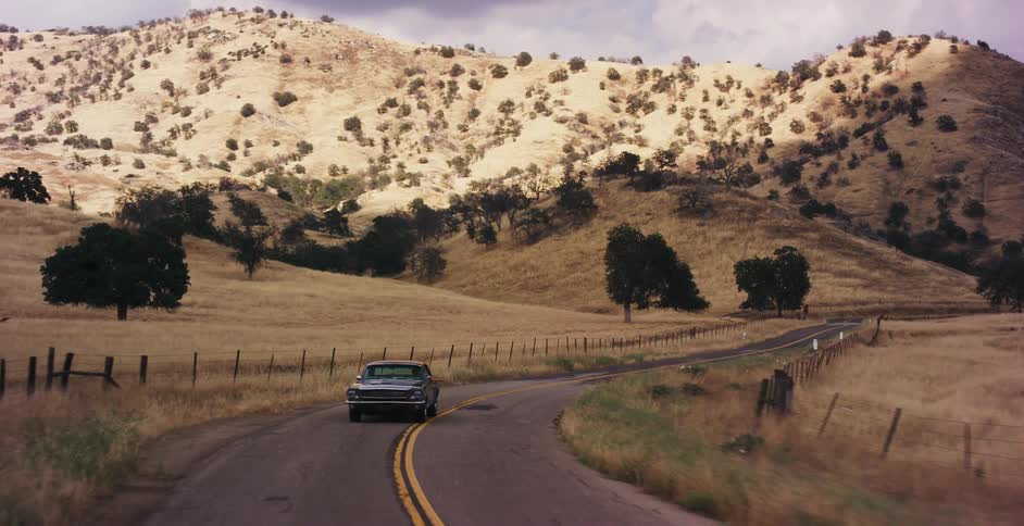 A vintage muscle car drives along a winding country road through rolling golden hills on a cloudy day
