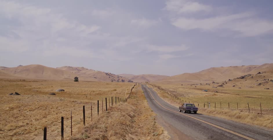A vintage muscle car drives down a long, lonely highway through the arid, golden hills of the countryside