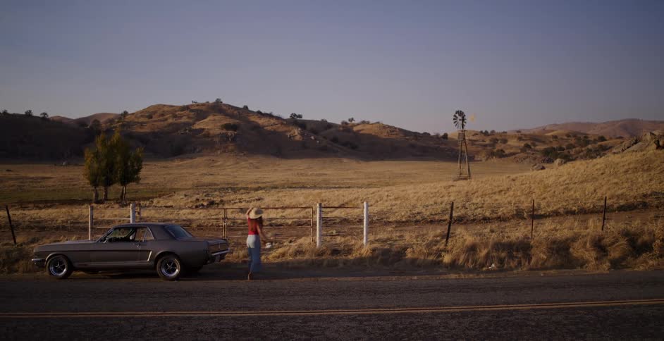 A woman driving a classic muscle car pulls over on a country road to get out and enjoy the scenic view of the rural landscape at sunset