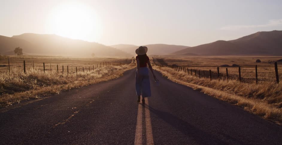 A young woman wearing a hat walks down a long, empty country road into the warm glow of the setting sun
