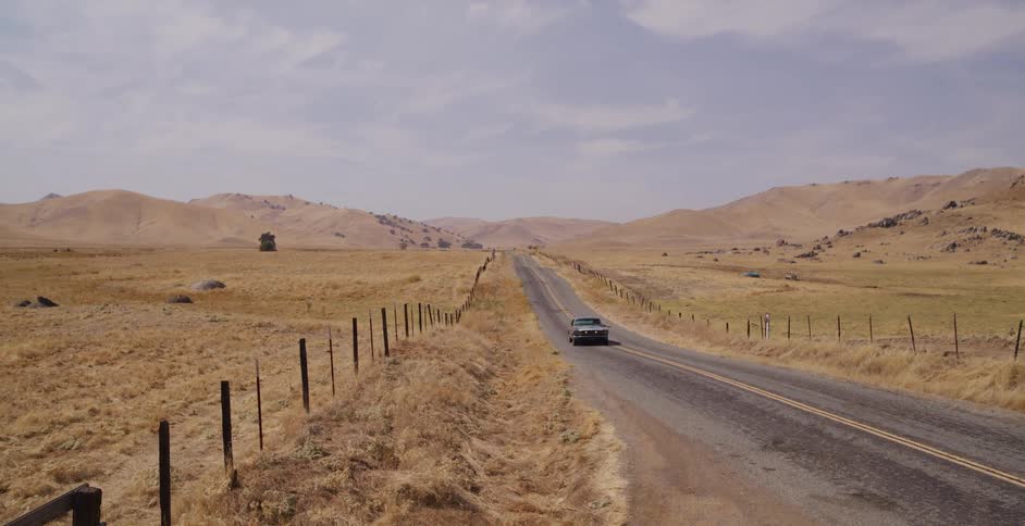 A vintage grey muscle car drives down a deserted, sun-bleached country road through rolling golden hills