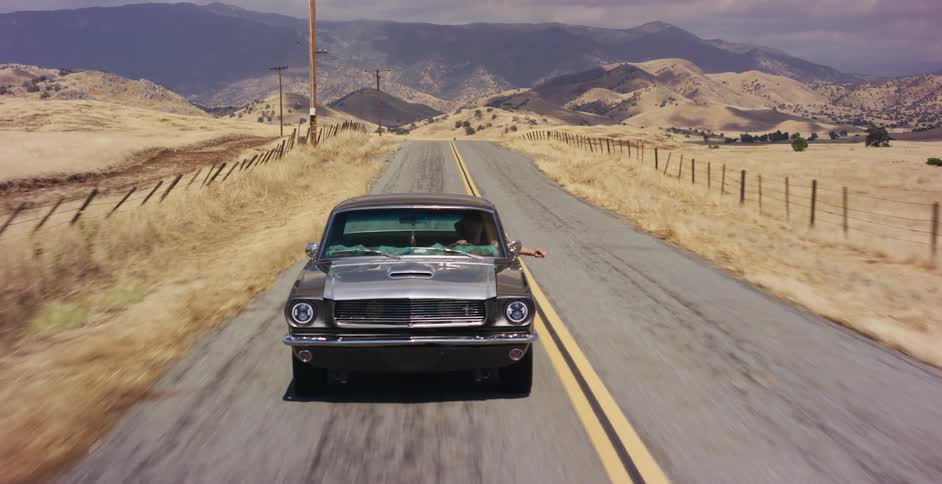 A classic muscle car drives down a deserted country road with rolling hills in the background on a sunny day