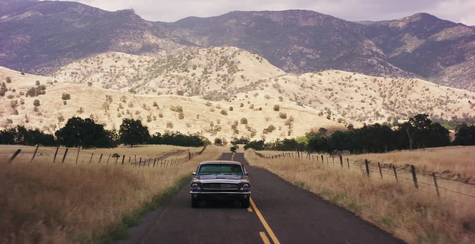 Aerial view of a classic black Mustang driving along a picturesque country road with golden fields and mountains