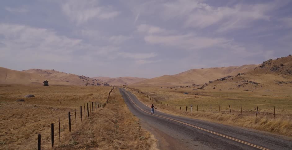 A carefree woman runs away from the camera down an empty country road through a vast, dry, hilly landscape
