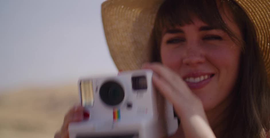 Close up of a smiling woman in a sun hat taking a picture with an instant camera and watching the photo develop in slow motion