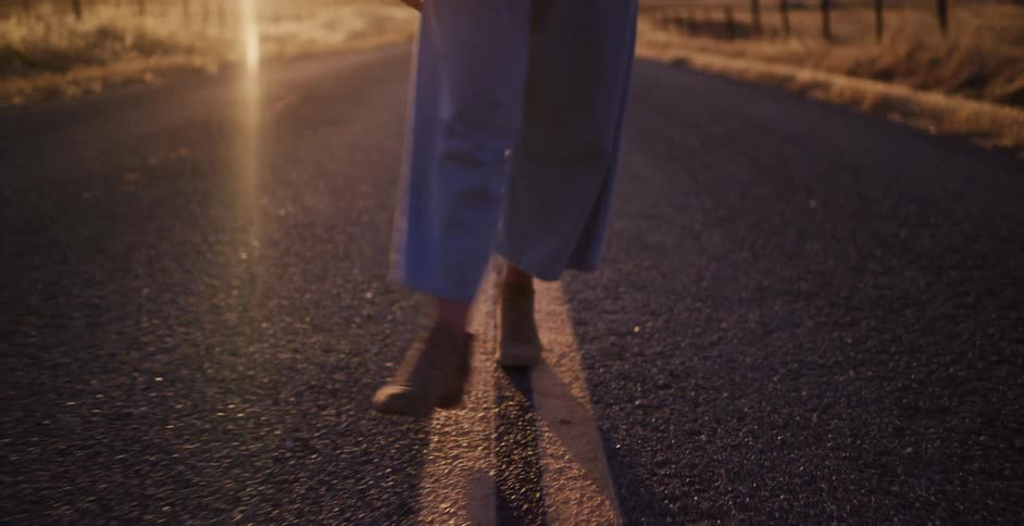 A joyful traveler wearing a hat and ankle boots strolls down a country road, smiling into the golden sunset