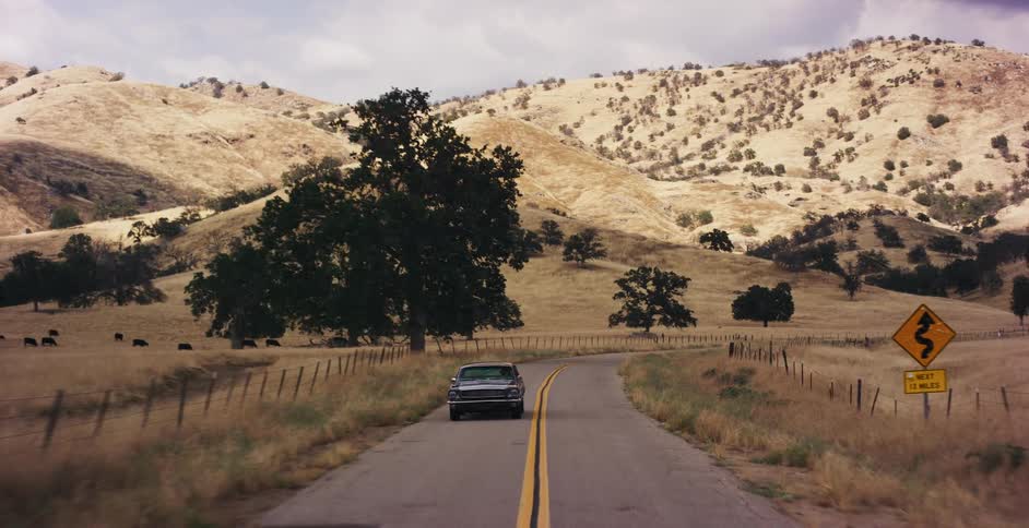 A classic car drives down a winding road through the golden hills of the countryside