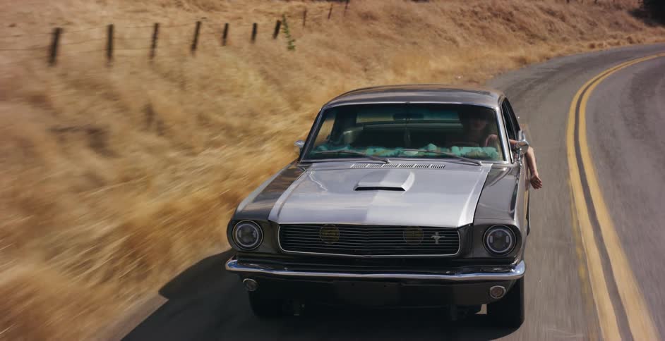 A classic silver Ford Mustang drives down a winding country road, with a passenger's arm hanging out the window in the summer sun