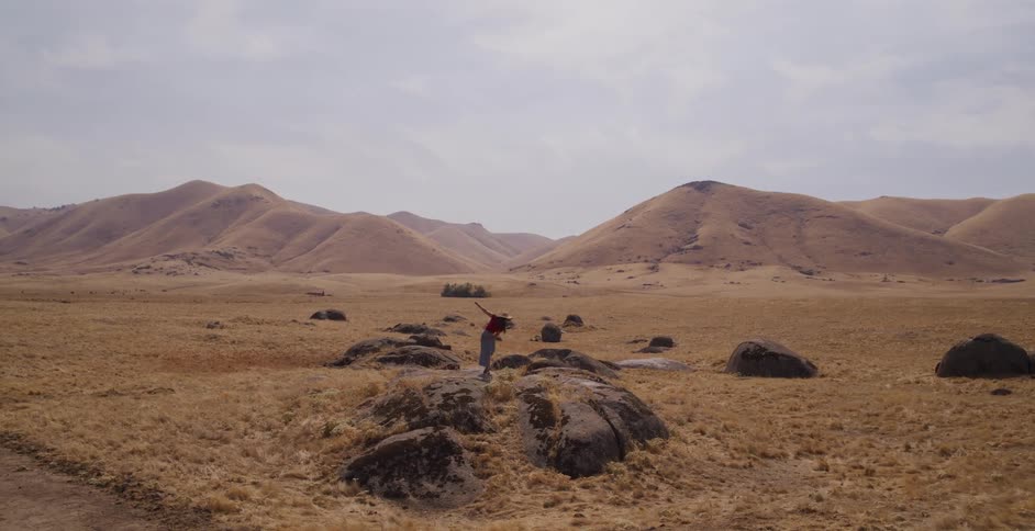 A woman with a hat joyfully runs through a barren, hilly landscape, climbs a large rock, and triumphantly stretches her arms out