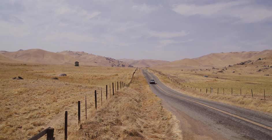 A classic muscle car drives down an empty country road through a dry, golden, hilly landscape